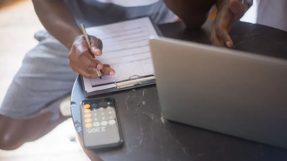A person filling out a form on a clipboard beside a smartphone and laptop