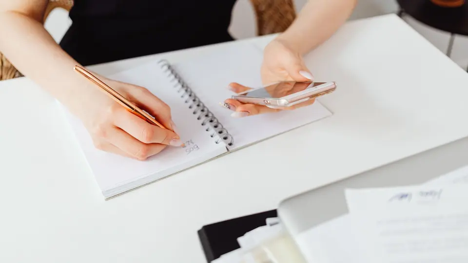 Hands writing notes in a notebook next to a smartphone and papers